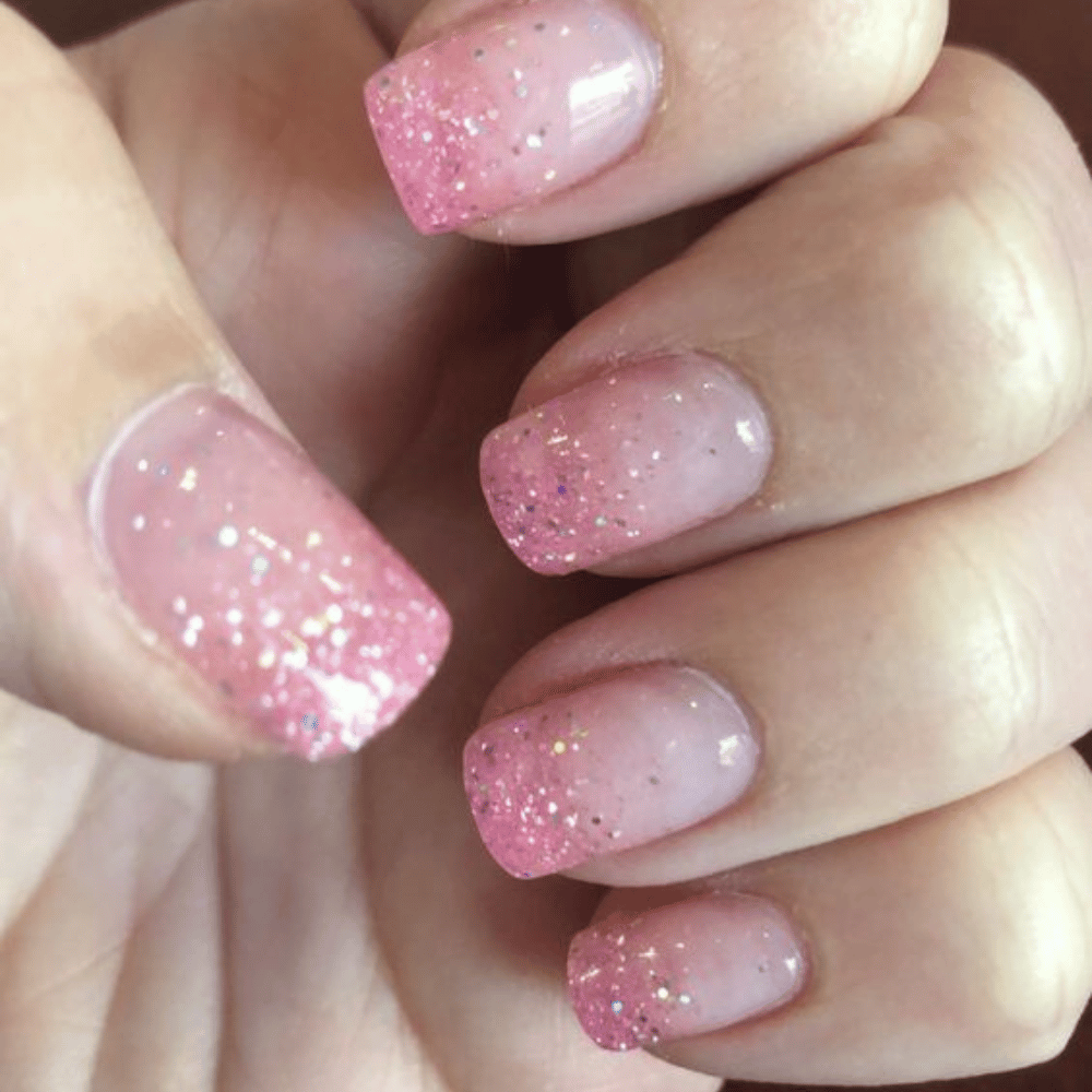 Hand with long, square-shaped nails featuring pink gradient from cuticle to tip, with glittery finish.