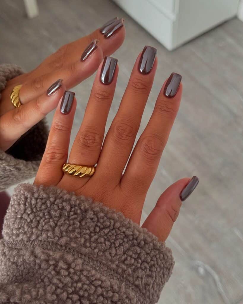 A hand with long, taupe polished nails and two gold rings, against a fuzzy beige sleeve backdrop.