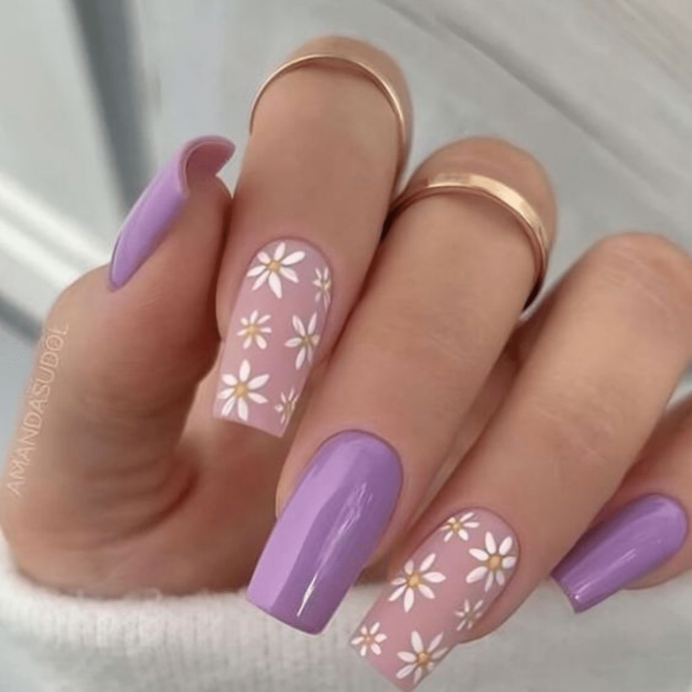 Close-up of hands with pastel purple nails featuring daisy and heart designs, wearing gold rings on fingers.