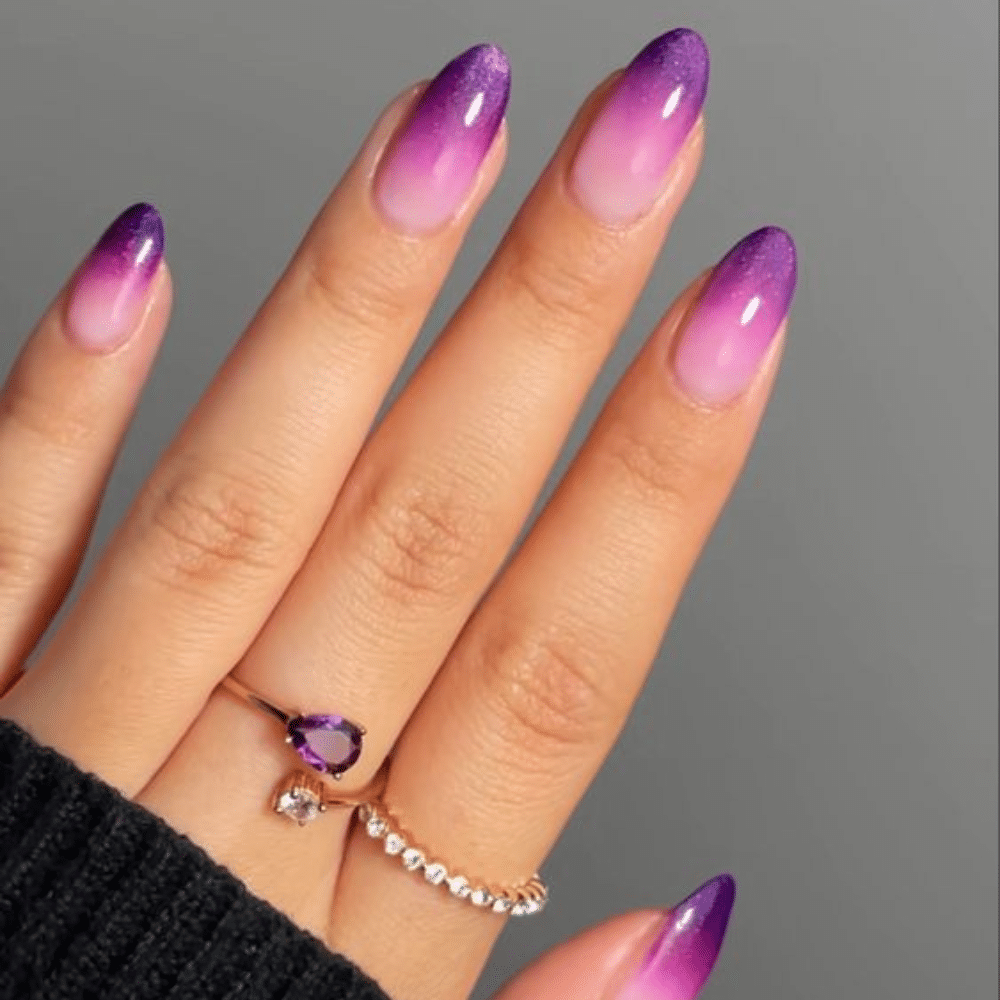 A hand with purple ombre nails and two rings, one with a purple gem and one with small diamonds.