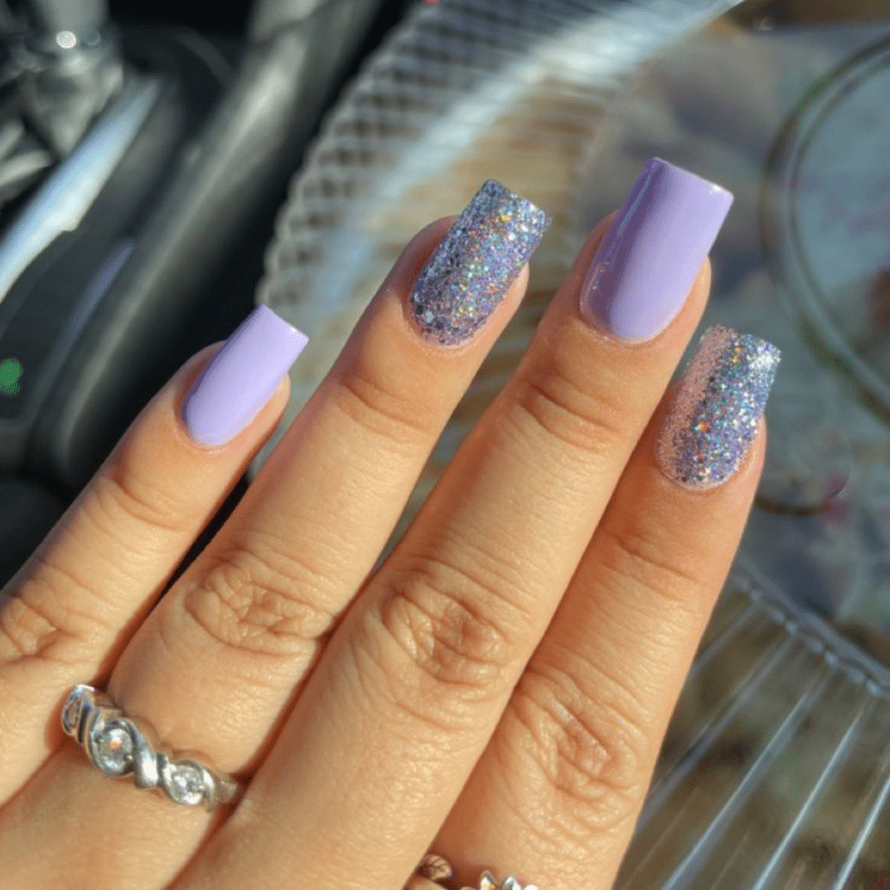 A hand with long, square-shaped nails painted in lavender and glittery silver polish, adorned with rings.