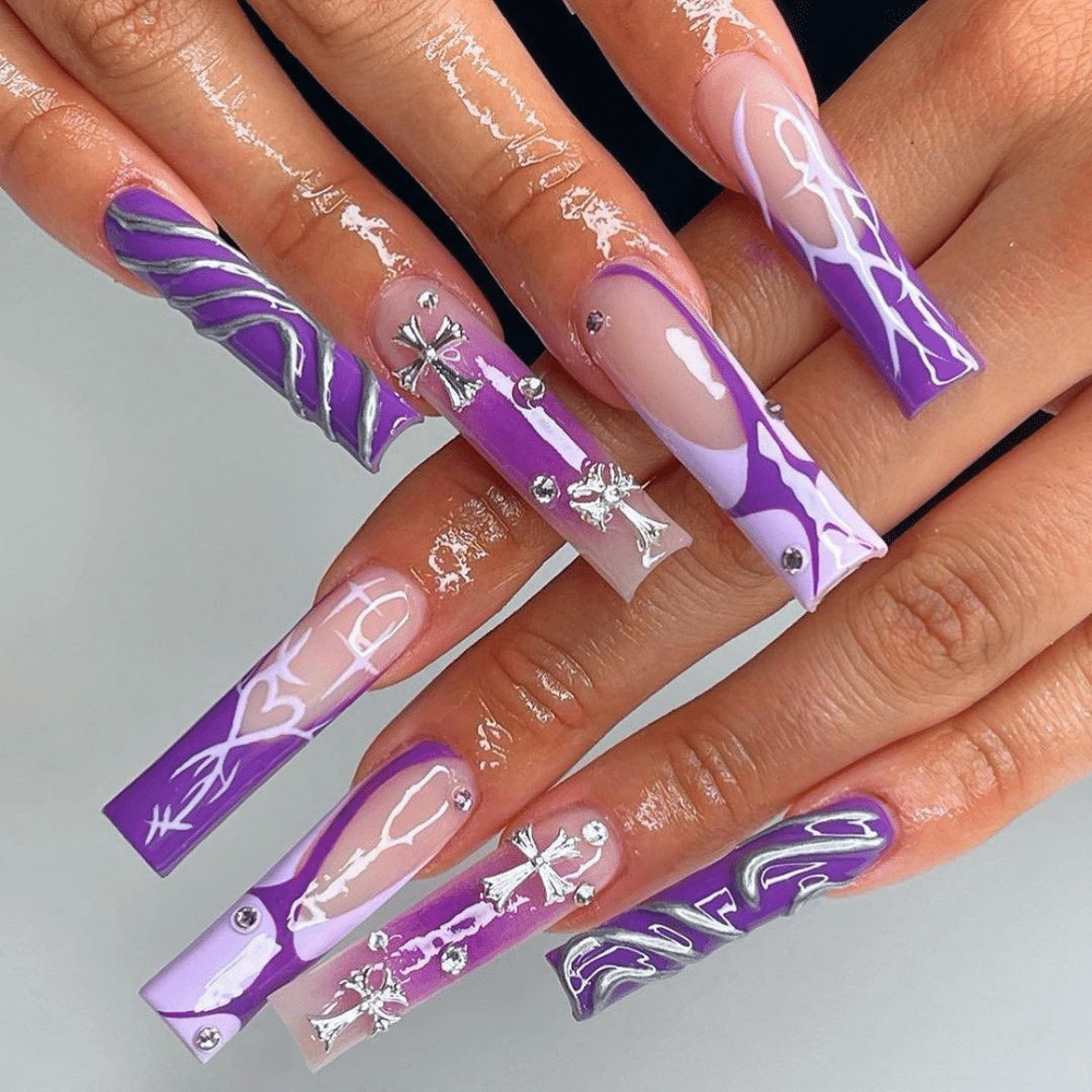 Hands with long, square-shaped nails featuring intricate purple and white designs with rhinestones and metallic accents.