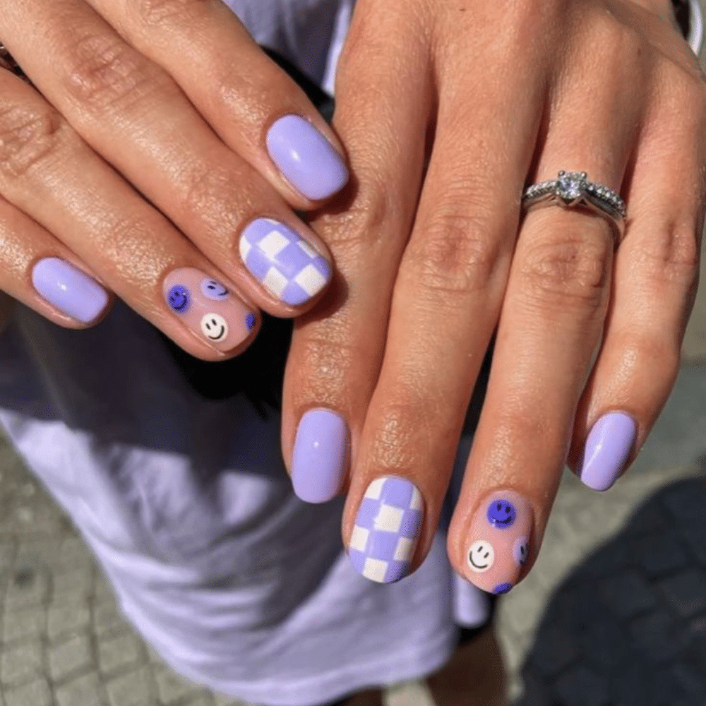 Close-up of hands with lavender nails, featuring checkered and smiley face designs, and a diamond ring.