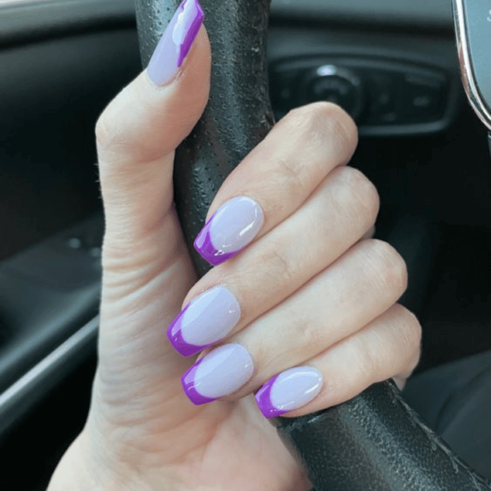 A hand holding a steering wheel, with light purple nails featuring darker purple French tips, against a car interior.