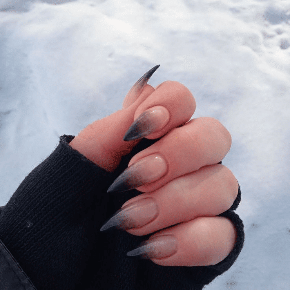 Hand with gradient nails transitioning from nude at the base to almost black at the tips, against a snowy background.