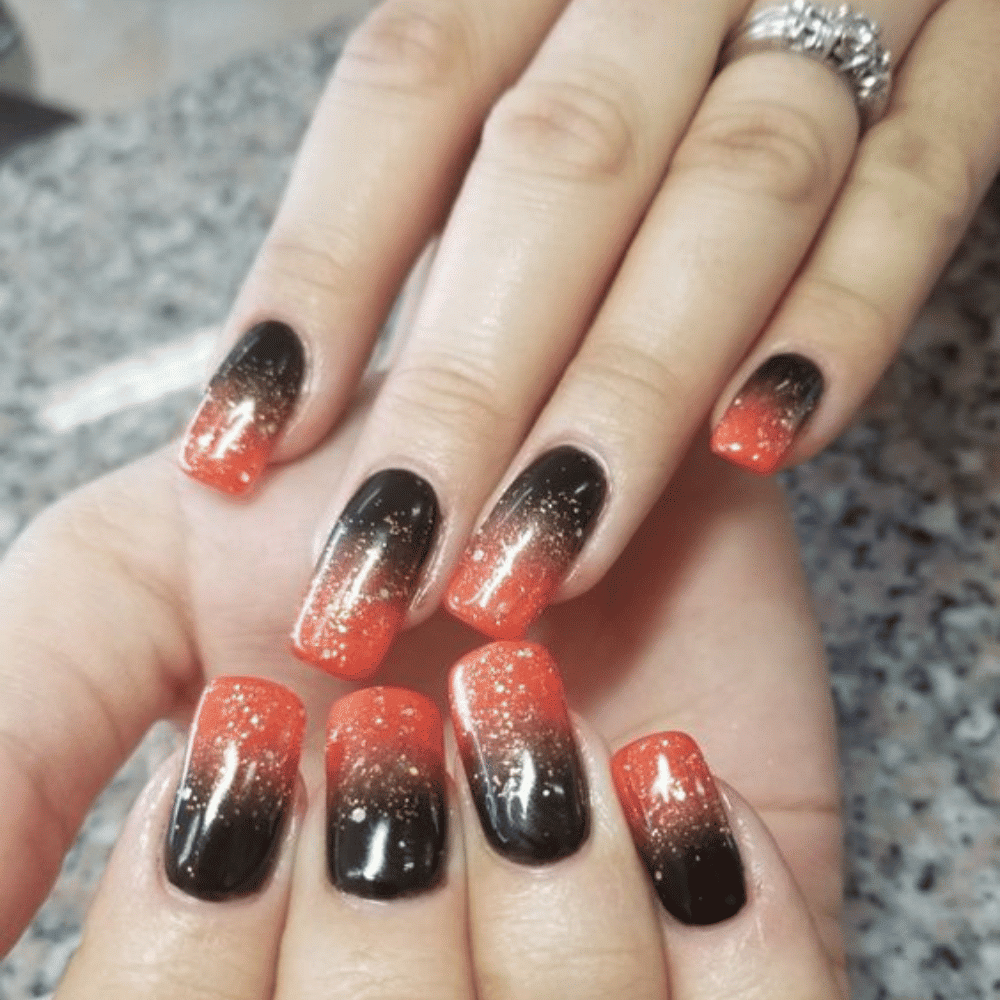 Close-up of hands with gradient nails transitioning from black at the base to bright orange tips, with a glittery finish.