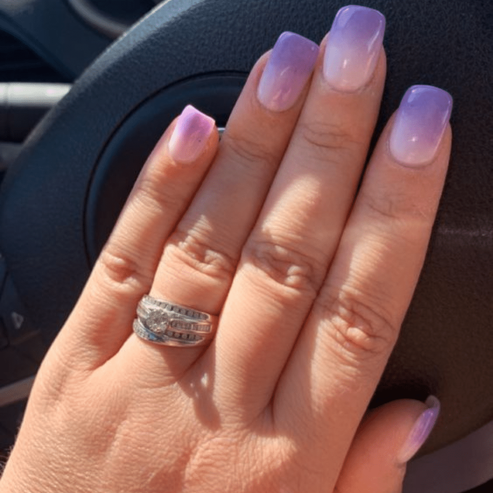 A hand with purple gradient nails and a diamond ring on the ring finger, against a car steering wheel.