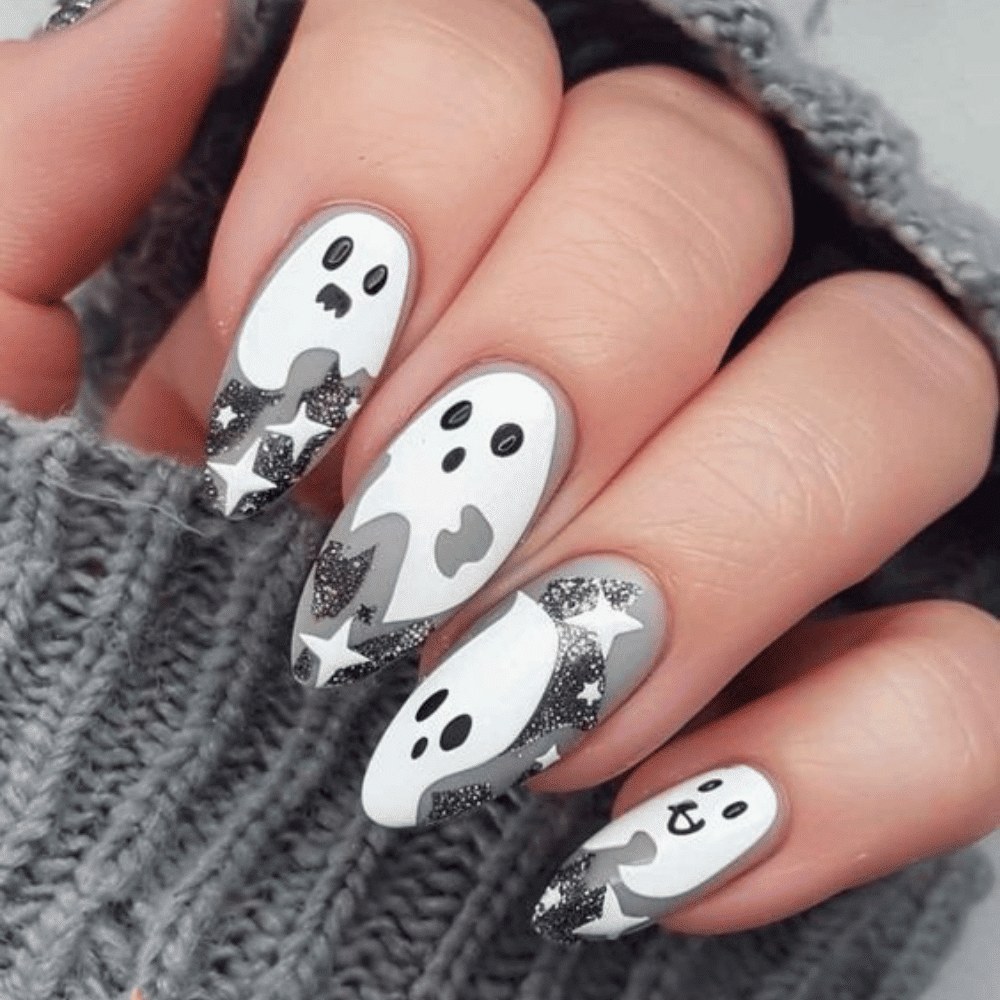 Close-up of a hand with long, almond-shaped nails painted white, featuring playful ghost faces and black glitter stars.