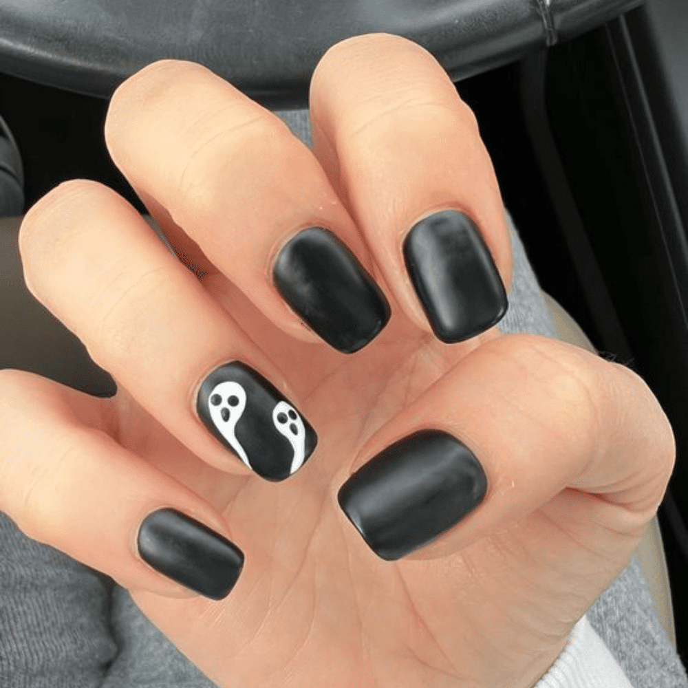 A hand with matte black nails, featuring two small white ghost figures with black eyes and mouths on the ring finger.