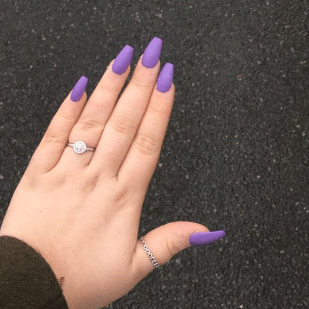 Close-up of a hand with long, purple, square-shaped nails and two rings, against a dark textured background.