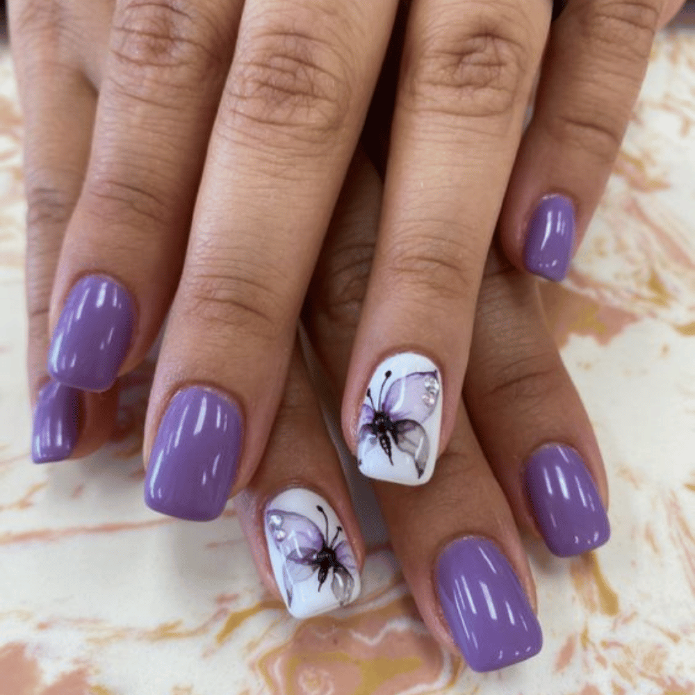 A hand with glossy purple nails and ring fingers featuring detailed butterfly designs with decorative rhinestones.