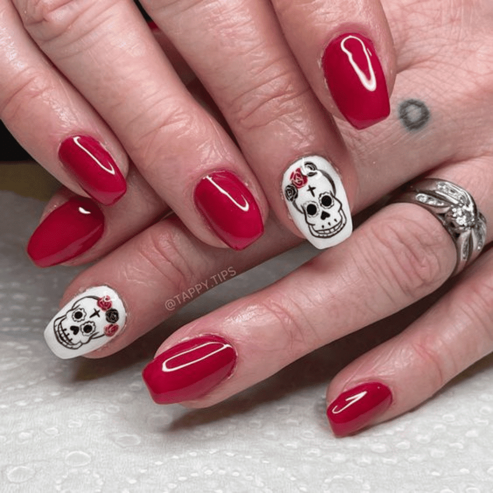 Hands with glossy red nails, except ring fingers which feature white base with detailed sugar skull designs.