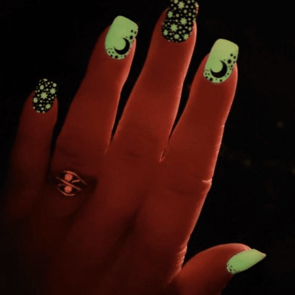 Hand with glow-in-the-dark nail art featuring crescent moons and stars, glowing green in the dark.