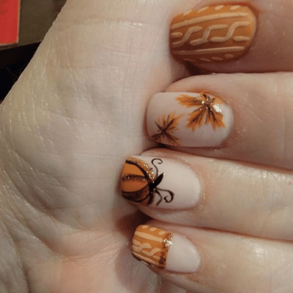 Close-up of hands with fall-themed nail art featuring orange, brown leaves, and pumpkin designs.