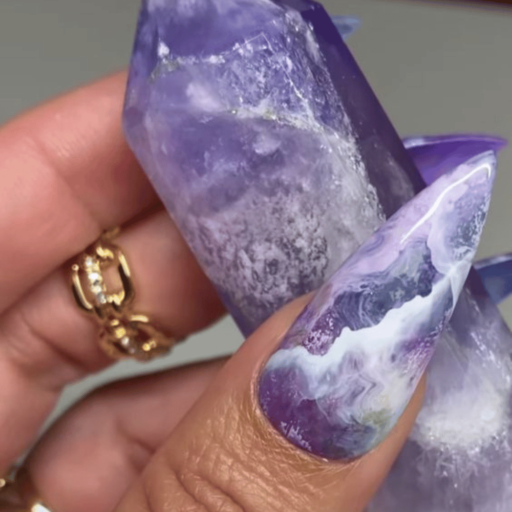 A hand holding a polished, purple crystal, with nails designed in a matching purple and white marbled pattern.