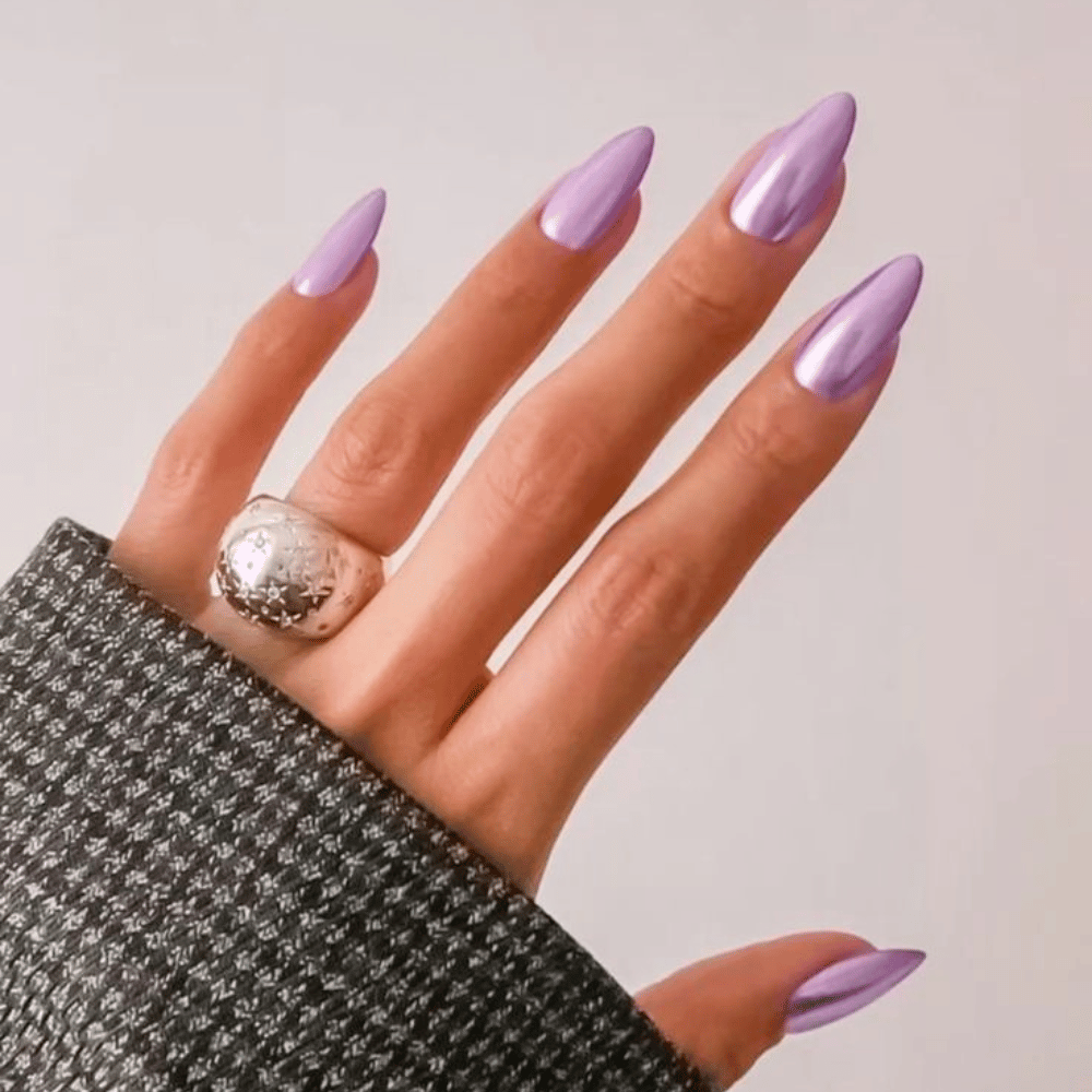 The image shows a close-up of a hand with long, pointed nails painted in a shiny lavender color and a large silver ring on the middle finger.