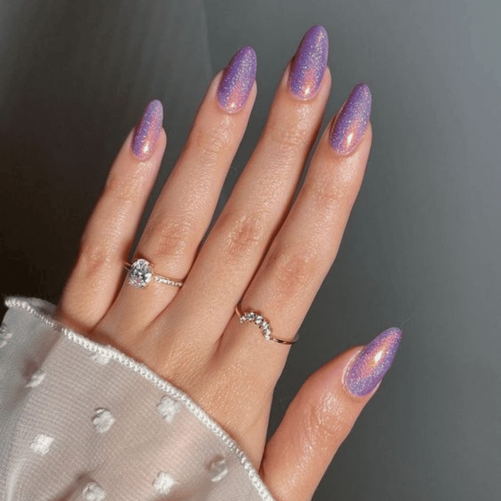 A close-up of a hand with long, almond-shaped nails painted glittery purple, adorned with two rings. One ring has a large, round gemstone, the other has a wavy design with small stones. Sheer polka-dot sleeve visible.