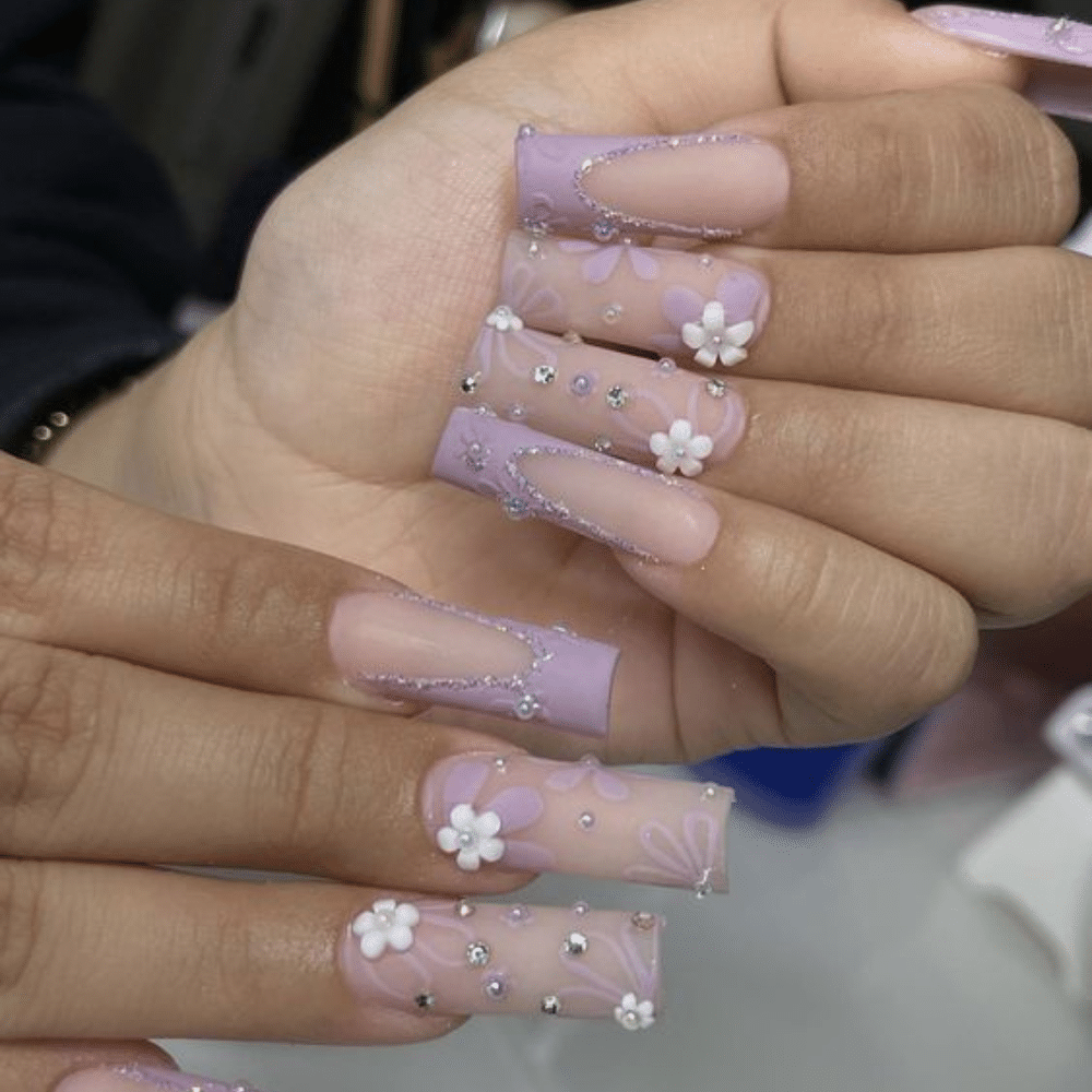 Close-up of a hand with long, square-shaped nails featuring a purple and nude design with 3D flowers and rhinestones.