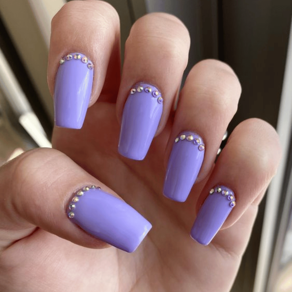 Close-up of long, square-shaped nails painted solid lavender, each adorned with small, round rhinestones near the cuticles in a semi-circle pattern.