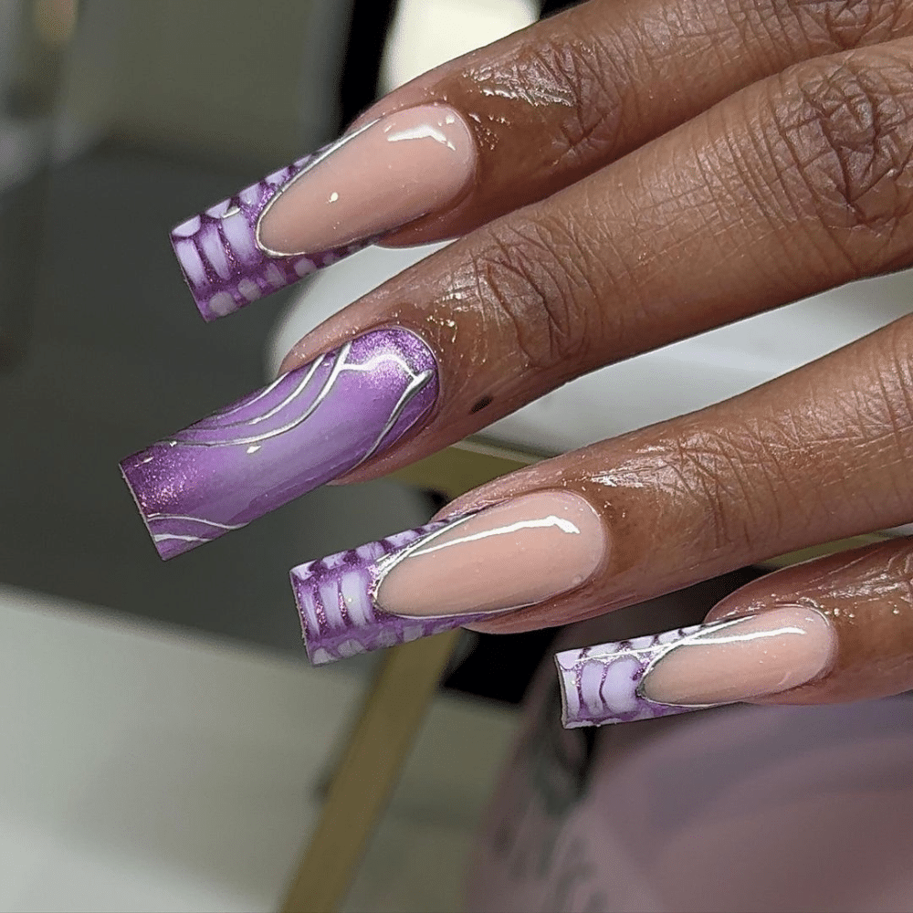 A hand with long, square-shaped nails featuring a purple and nude design with intricate patterns.